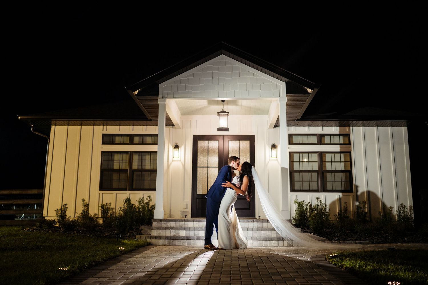 A couple shares a romantic moment at night outside a white barn venue with dramatic lighting and shadows.