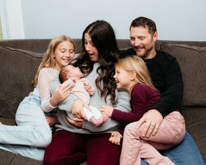 A family cuddles together on a grey couch while holding a newborn baby. Family newborn in home session. Newborn photographer.