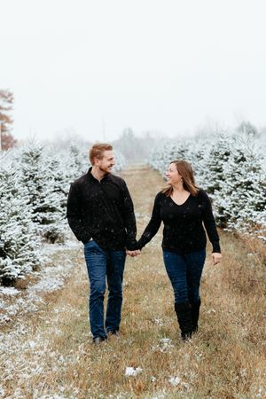 Winter engagement photos of a couple walking through a snowy Christmas tree farm. Central Michigan Christmas Tree Farm Photographer.