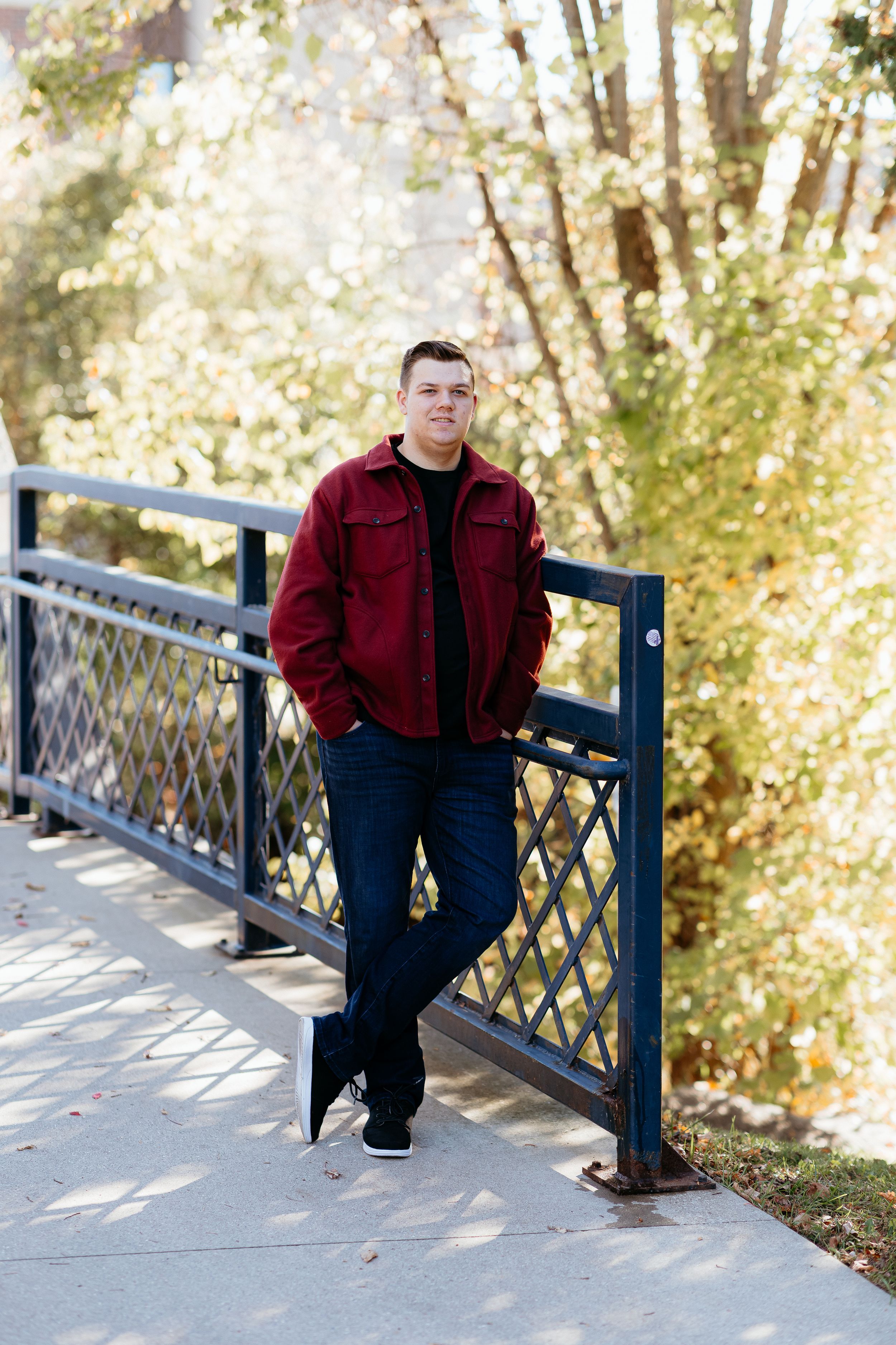 A Senior boy in a burgundy jacket poses candidly on the blue bridge in island park mount pleasant Michigan in fall. A Senior Photographer.