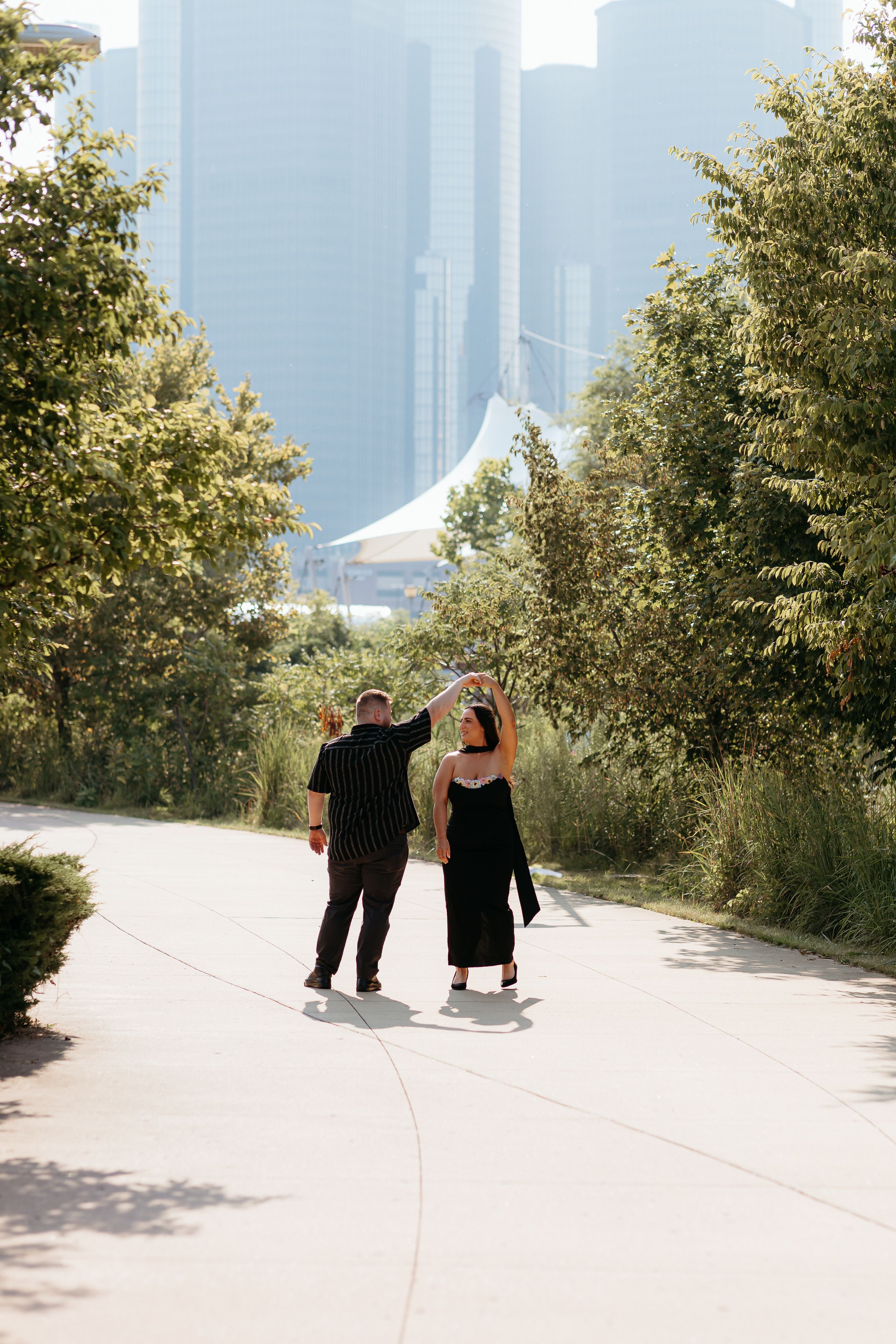 Couple walking together on a path with the Detroit city skyline visible in the background. Waterfront Detroit Michigan. Engagement Session.