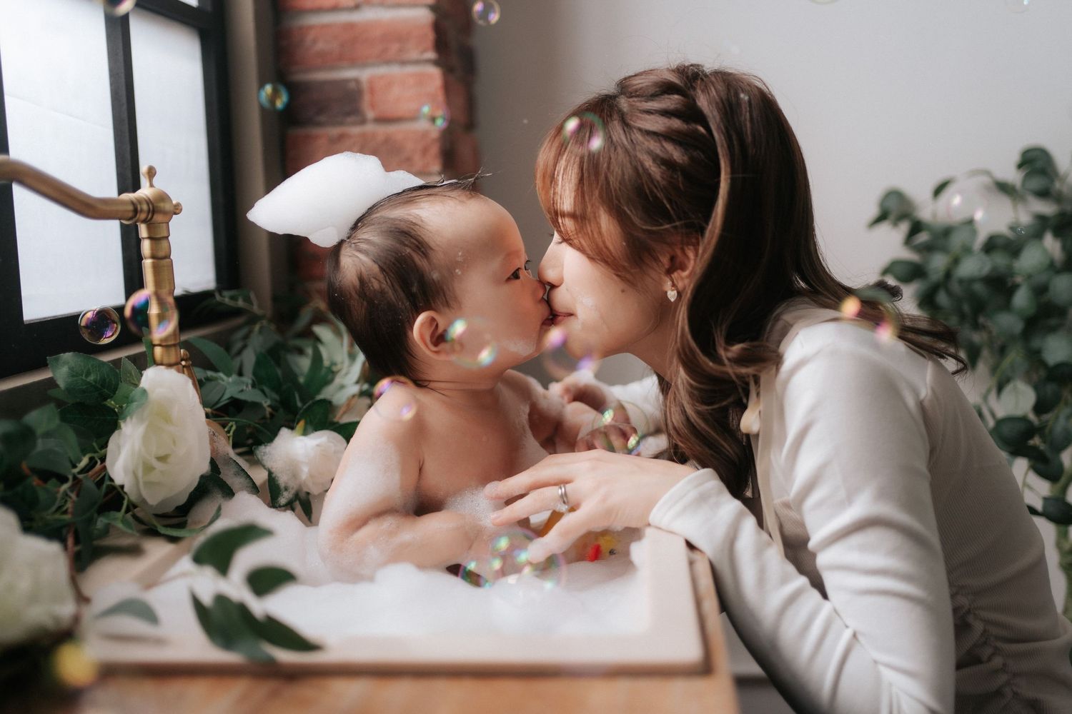 A loving mother tenderly kisses her baby during a bubble bath, surrounded by white flowers and greenery一位慈愛的母親在泡泡浴中溫柔地親吻著她的寶寶，周圍環繞著白色的花朵和綠植。