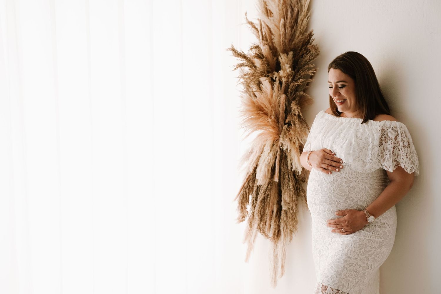 Smiling pregnant woman in white lace dress holding belly beside pampas grass decor.