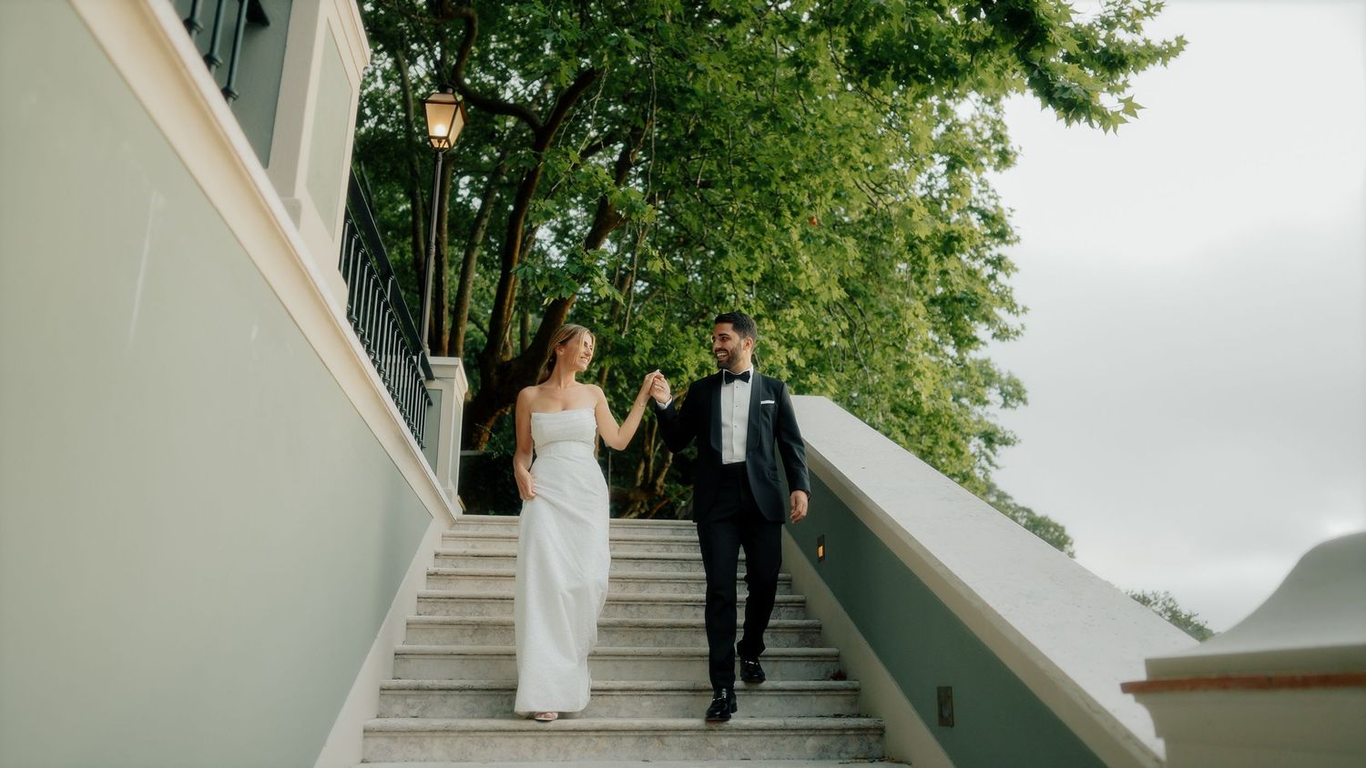 A couple in wedding attire descend white outdoor stairs together while holding hands on their special day.