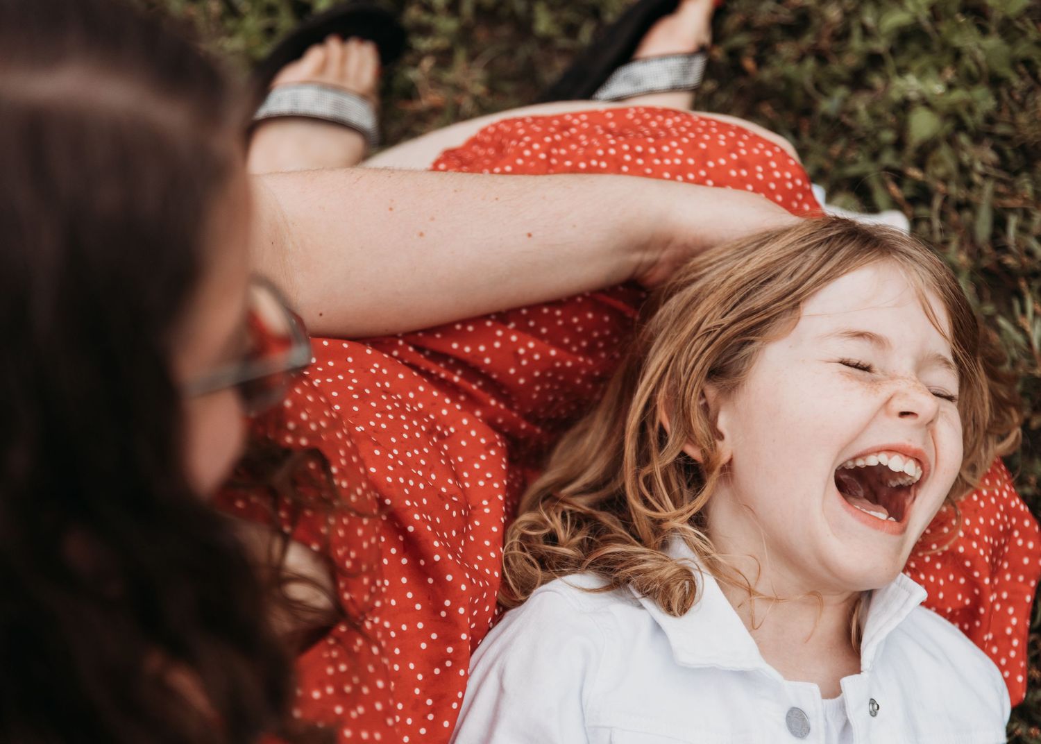 Two people share a joyful moment laying on a red polka dot blanket outdoors in the grass.