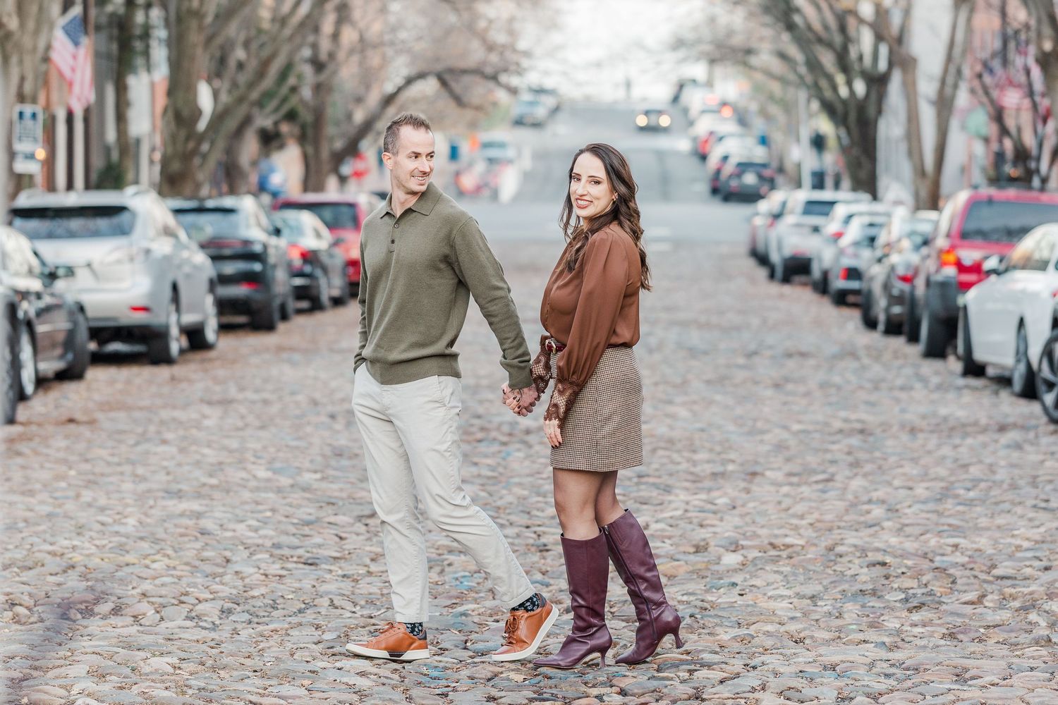 People walking on a cobblestone street lined with parked cars and autumn leaves on a cloudy day.