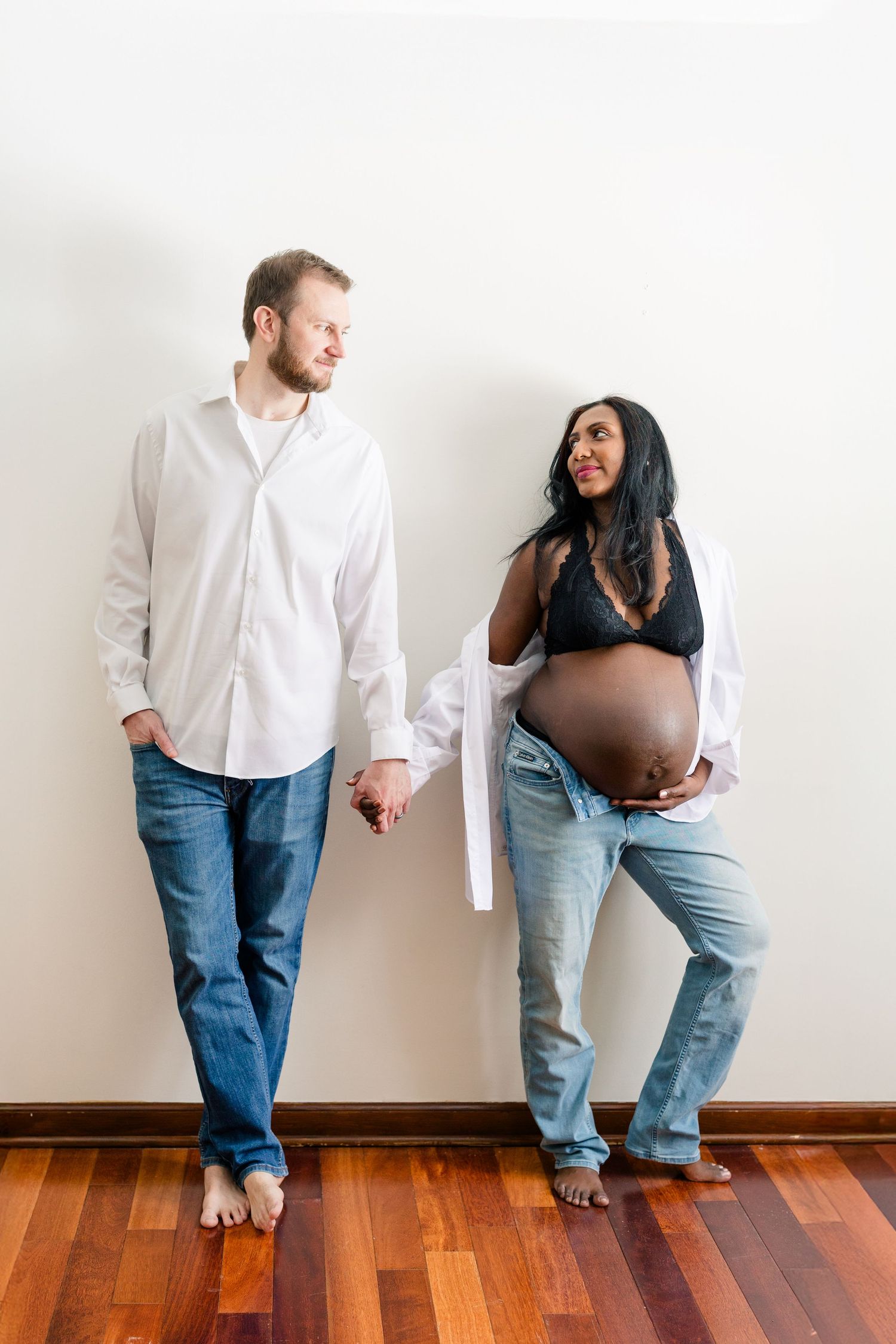 An expectant couple stands barefoot on hardwood floors against a white wall, holding hands and looking at each other.