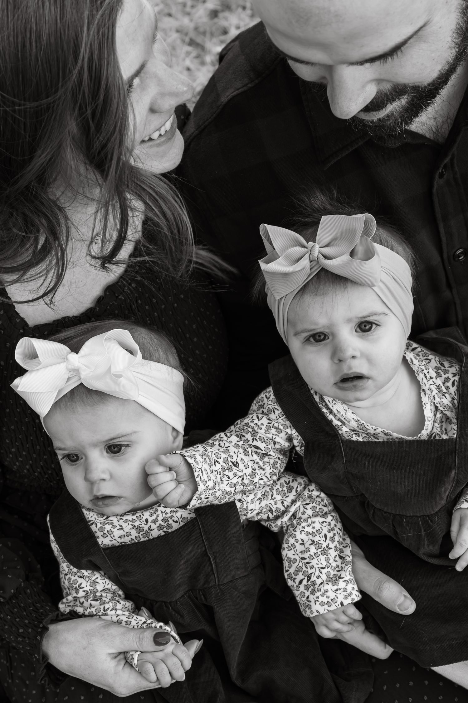 Black and white photo series showing baby twin girls wearing a floral dress and bow headband from different angles with mother and father.