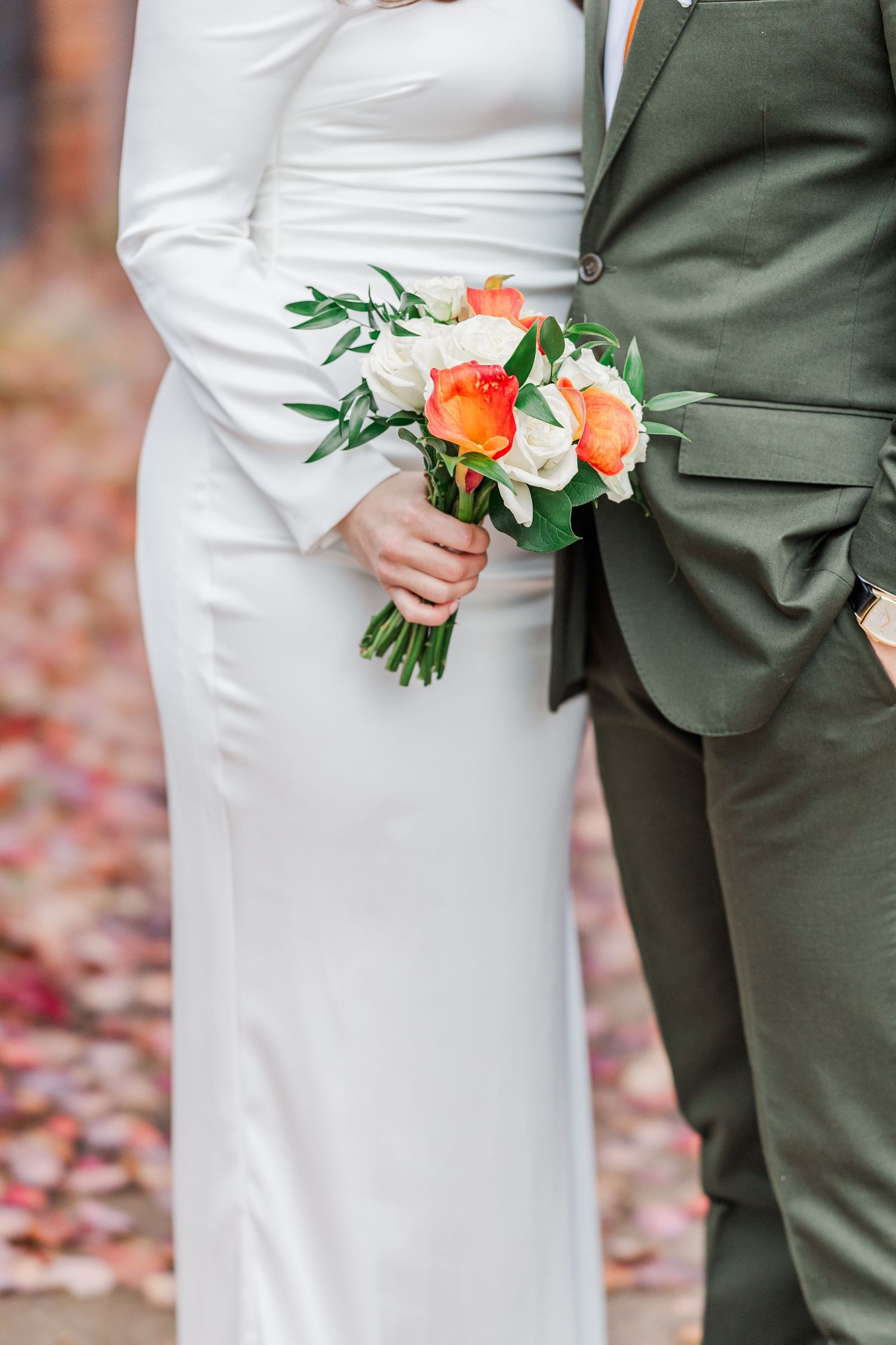 A couple poses for wedding photos beneath vibrant red autumn trees with fallen leaves scattered on the ground.