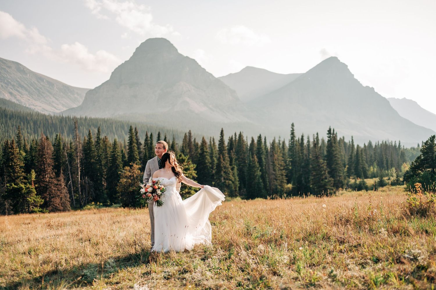 A couple in wedding attire embraces in a scenic mountain meadow surrounded by evergreen forests and majestic peaks.