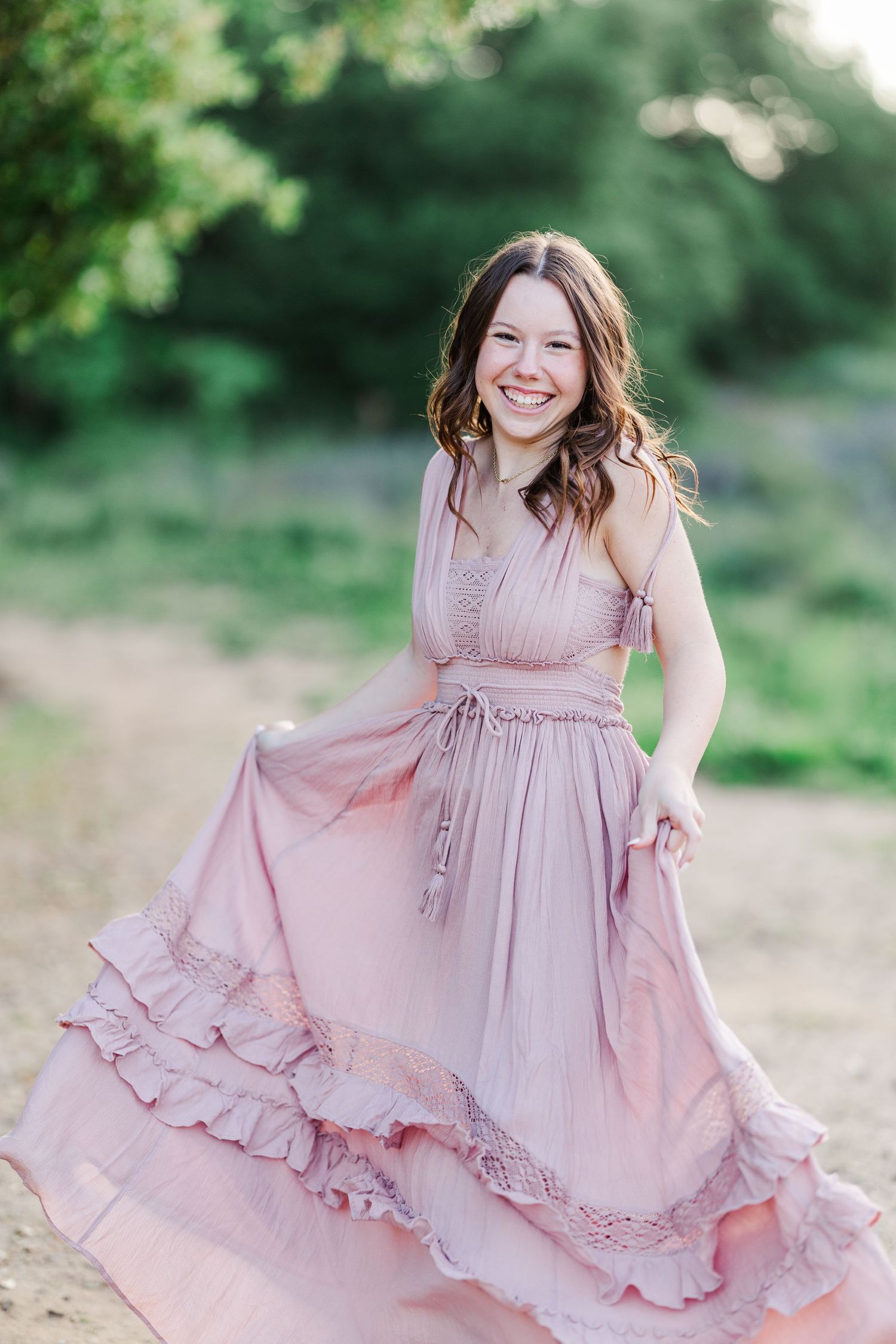 A person in a flowing dusty rose maxi dress twirls joyfully in a sunlit outdoor setting with green foliage.