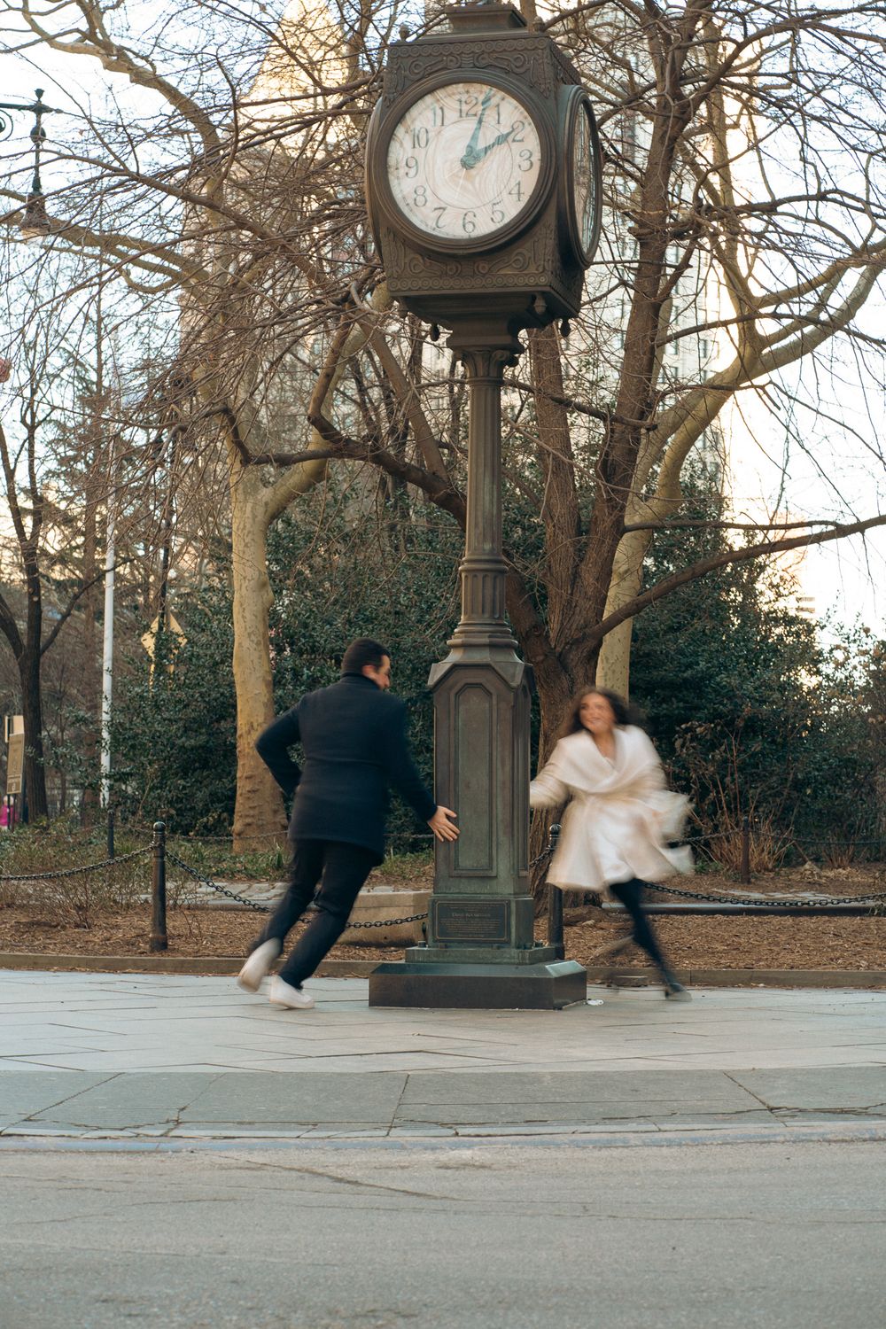 Two figures playfully circling around a vintage clock post in a park during winter.