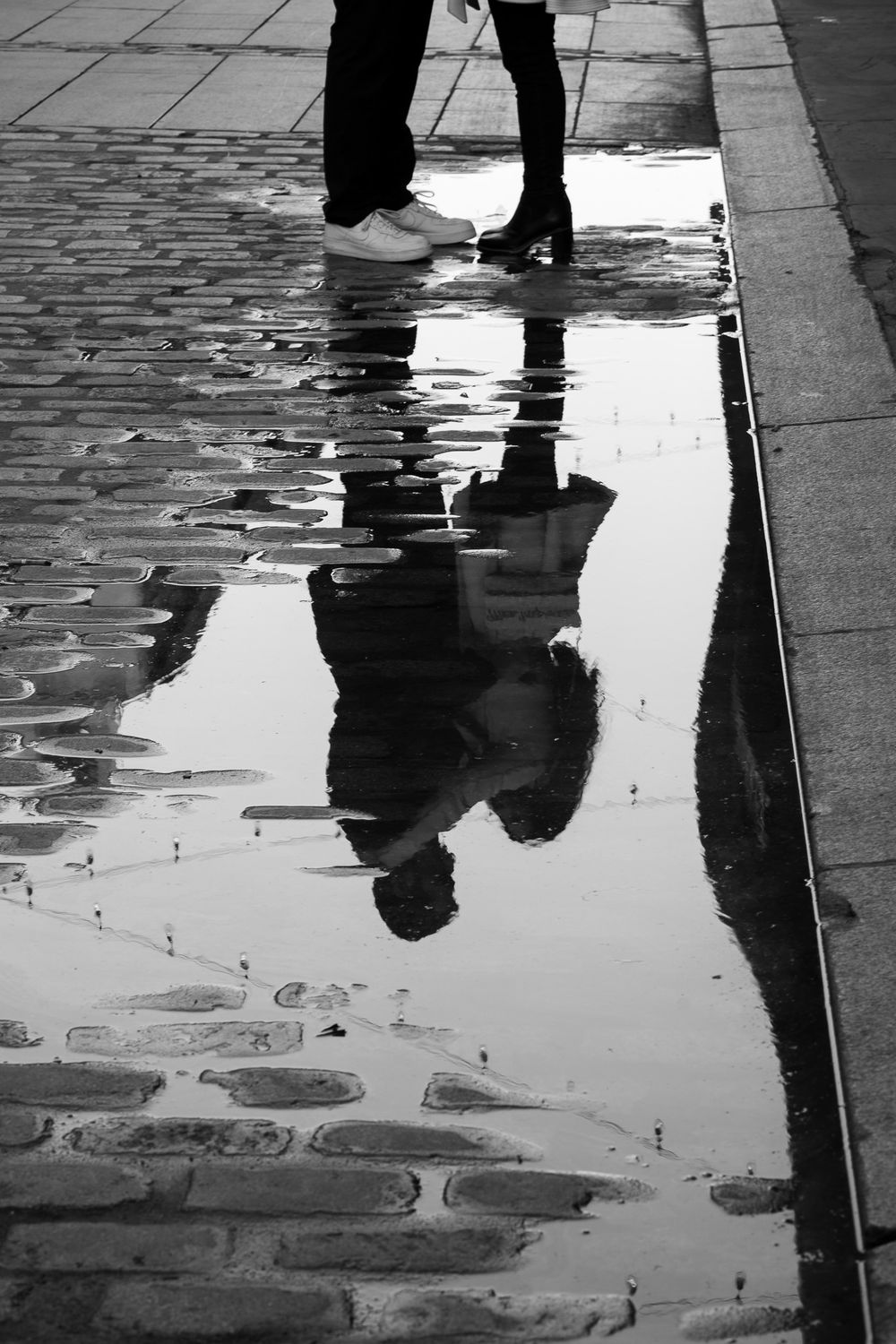 Black and white reflection of people walking on wet cobblestone street creates dramatic shadows and silhouettes.