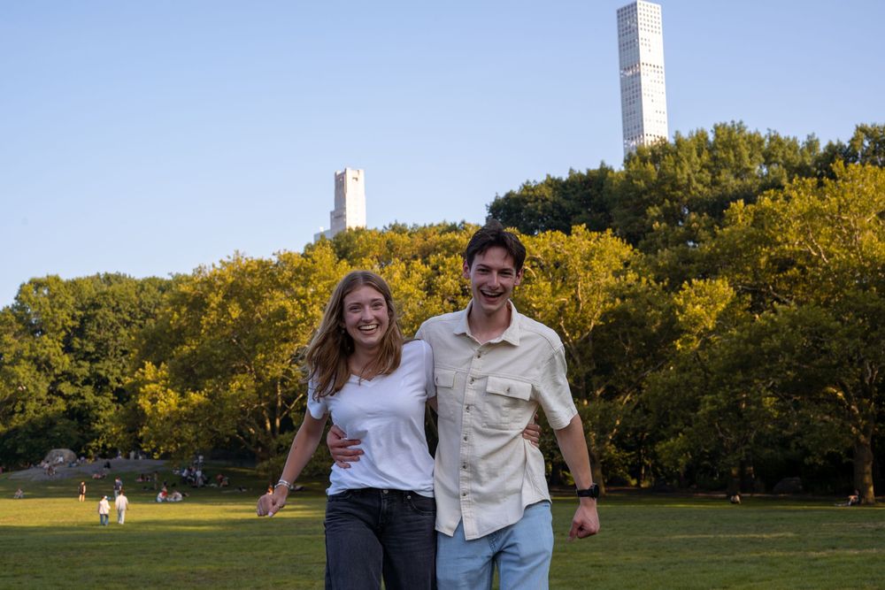 A couple walks together on a grassy field during golden hour with trees in the background.