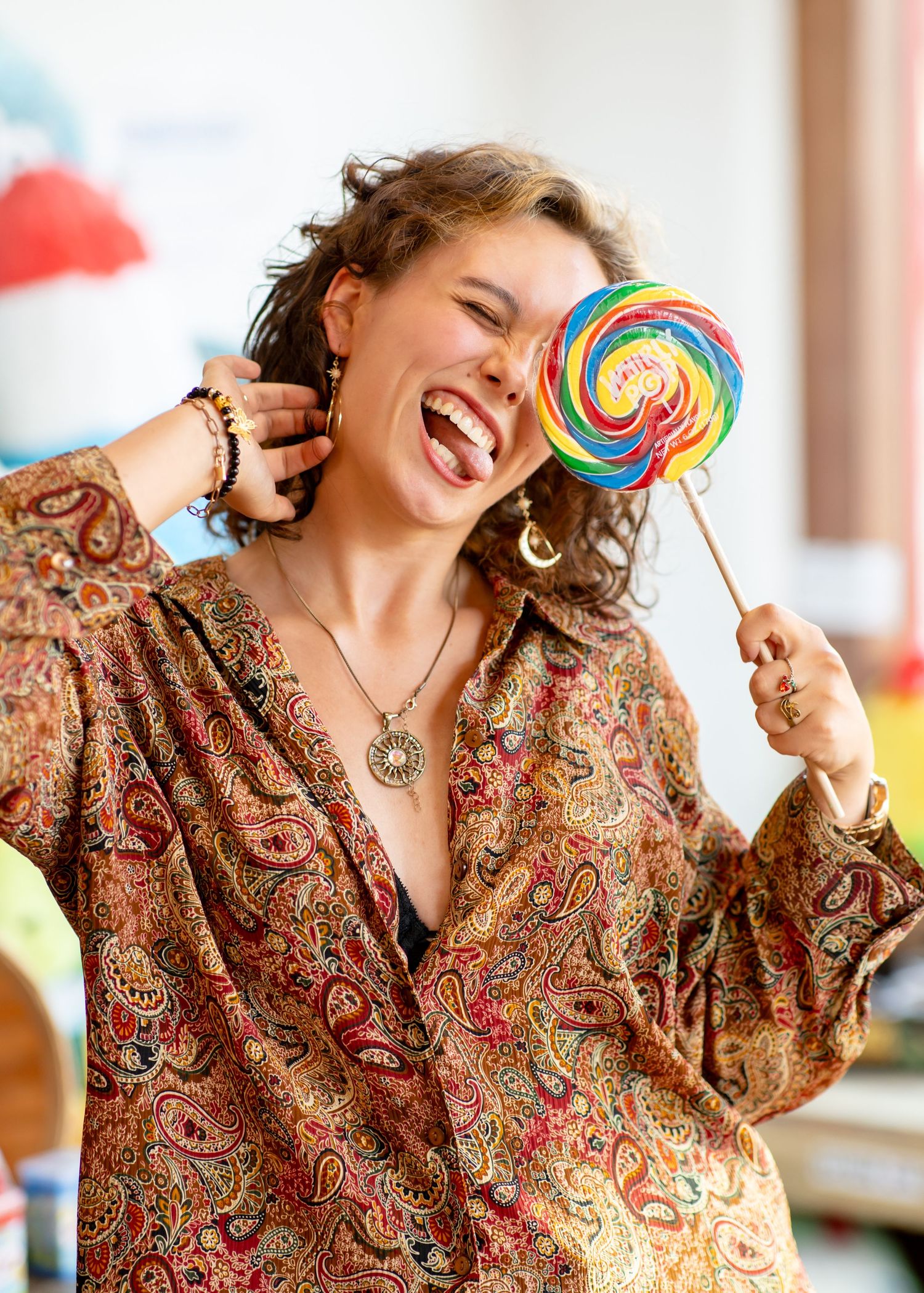 Joyful person in a patterned paisley dress playfully posing with a colorful rainbow lollipop.