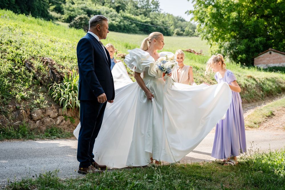Wedding party members help adjust a bride's flowing white dress on a sunny outdoor path.
