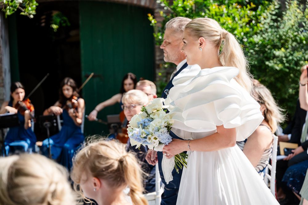 A wedding ceremony in progress with attendees gathered outdoors under green foliage.