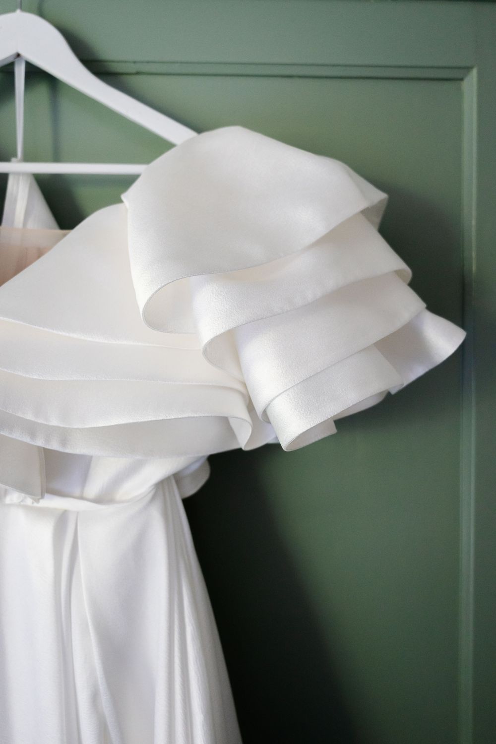 Close-up detail of a white wedding dress shoulder featuring elegant layered ruffles against sage green wall.