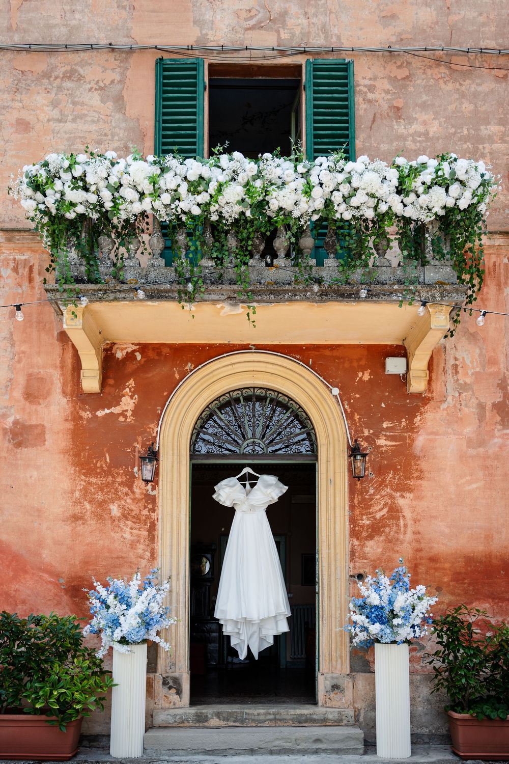 White wedding dress hangs in doorway beneath floral-adorned balcony with green shutters.