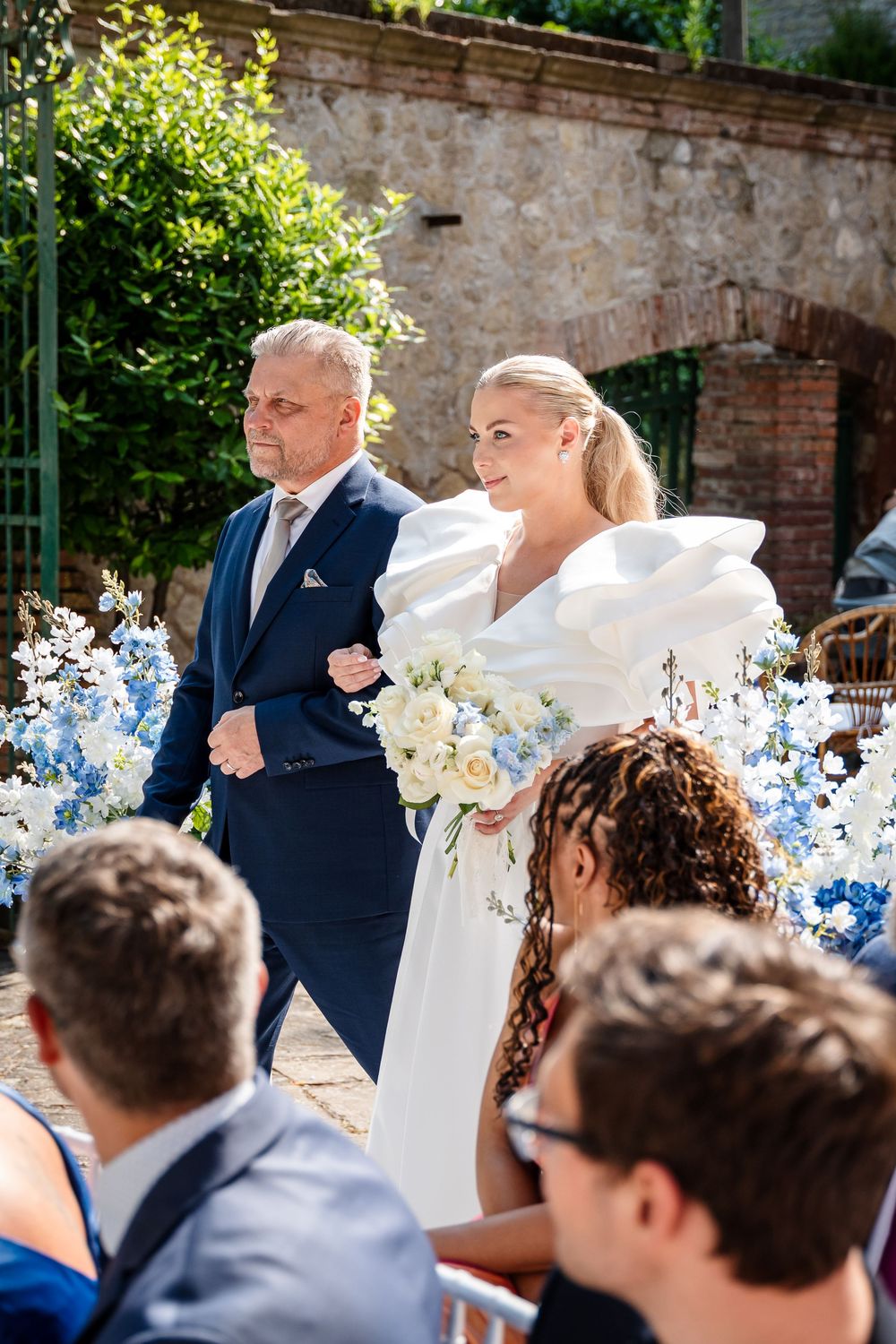 Wedding party celebrates outdoors with blue and white floral decorations against stone walls.