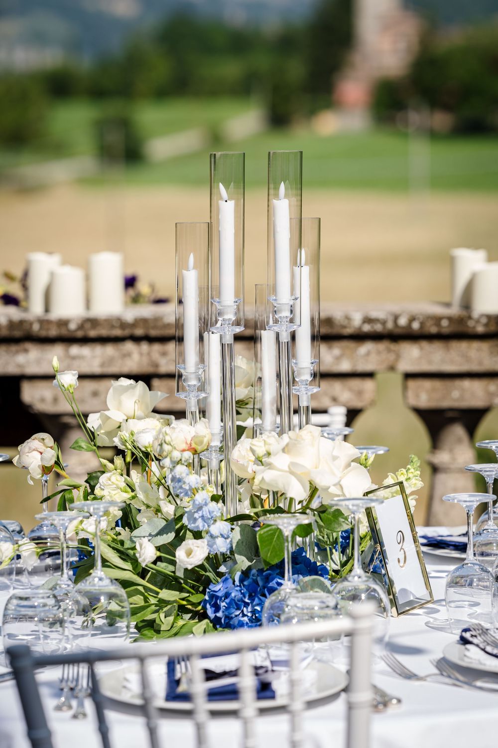 Wedding reception table decorated with blue hydrangeas, white roses, and tall glass candleholders.