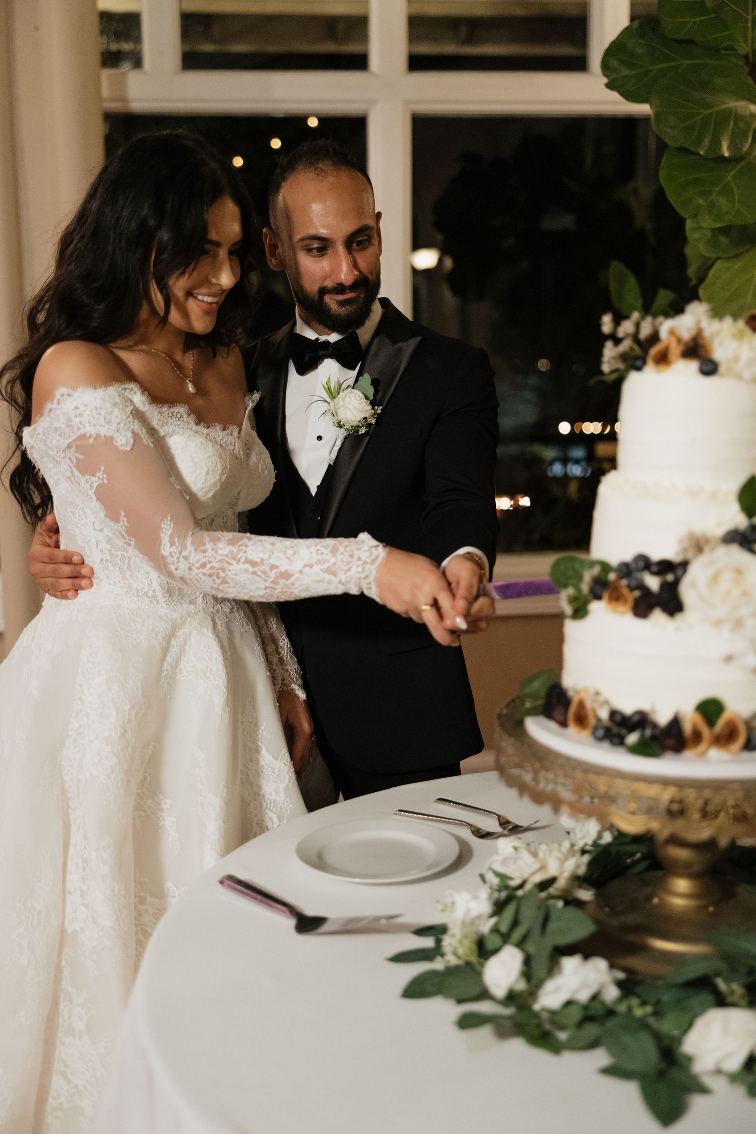 A couple cutting their elegant white wedding cake decorated with greenery at their reception.