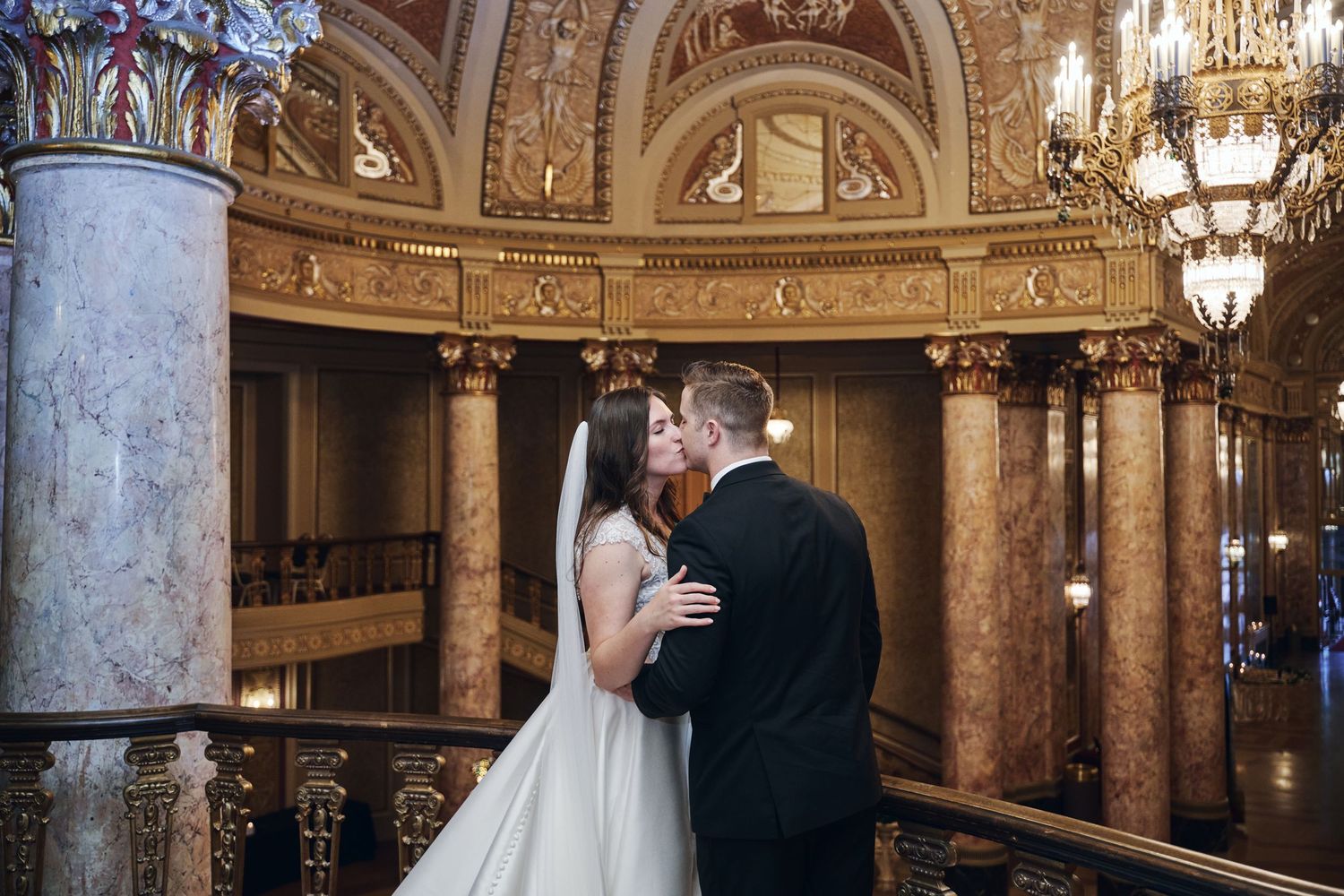 A couple shares an intimate moment in an ornate ballroom with marble columns and crystal chandeliers at the Rialto Square Theatre.