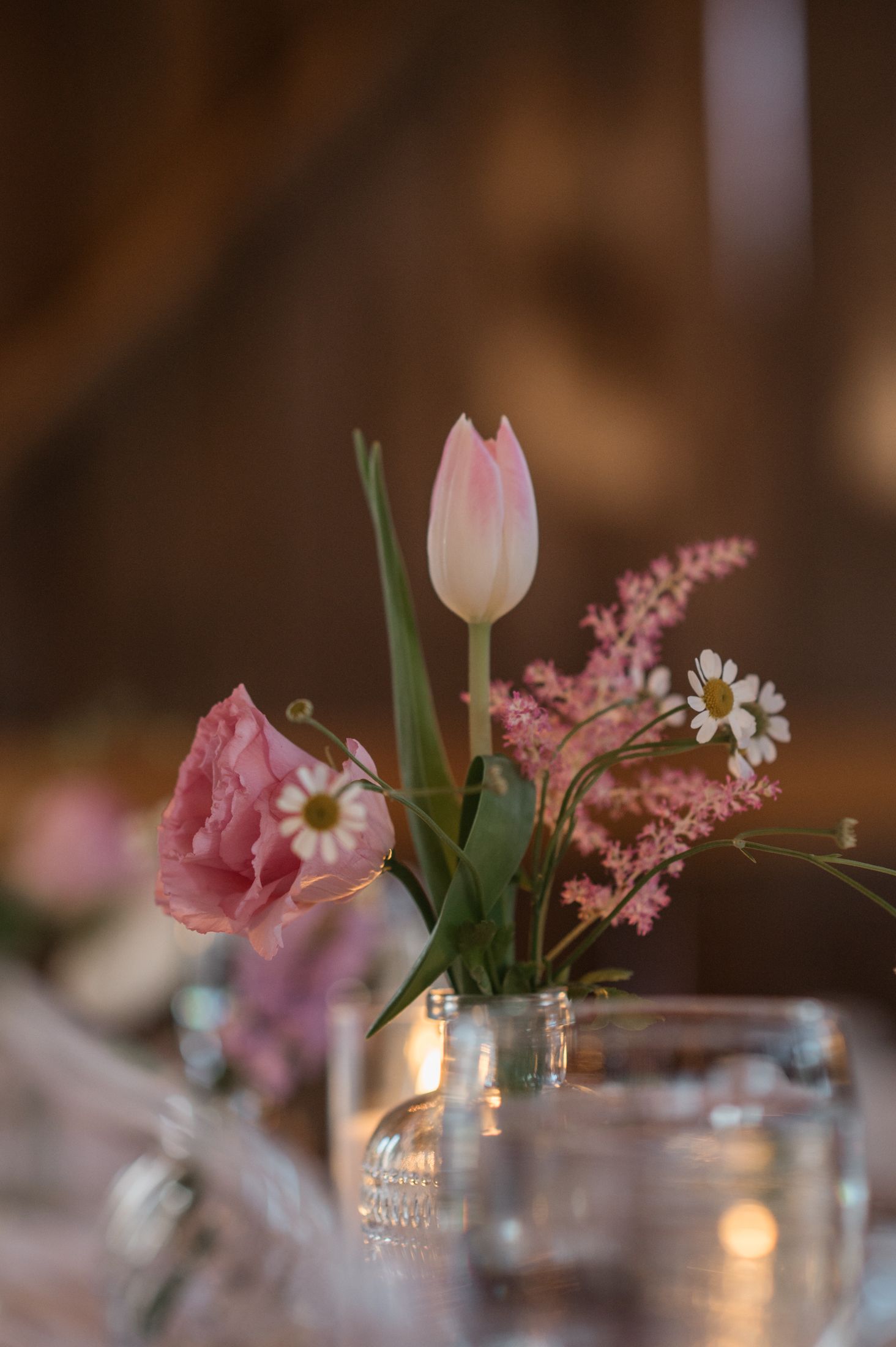 A delicate arrangement of pink tulips and daisies in a glass vase with glowing candlelight in the background.