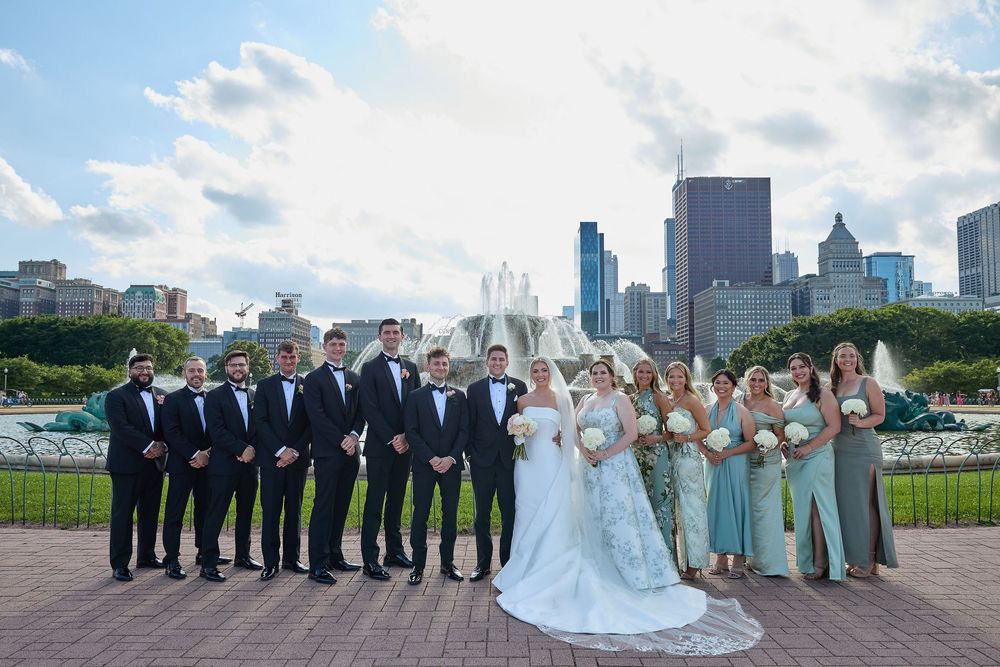 A wedding party poses together against the Chicago skyline at a park on a sunny day with blue skies and white clouds.