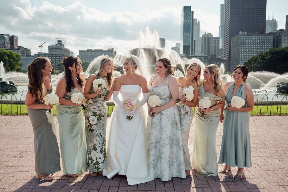 Bridal party in sage green dresses and white gown pose together at outdoor venue with city skyline in background.