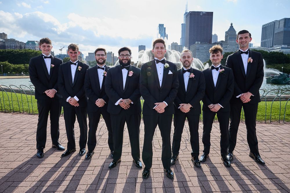 A group of groomsmen in black tuxedos pose together outdoors for a formal wedding photo.