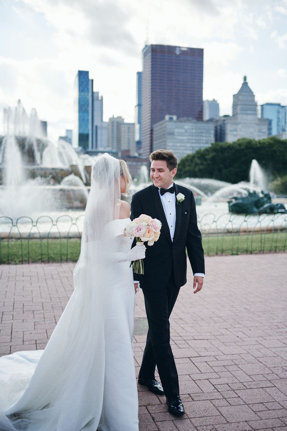 A wedding couple walks near Buckingham Fountain with the Chicago skyline and fountains in the background.