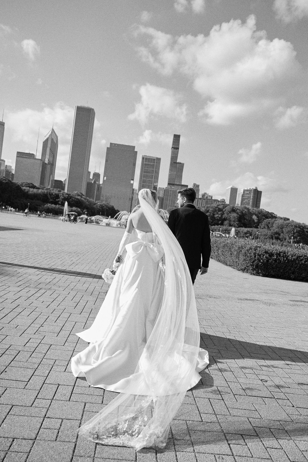Black and white photo of newlyweds walking along brick pavement with Chicago's urban skyline in the distance.