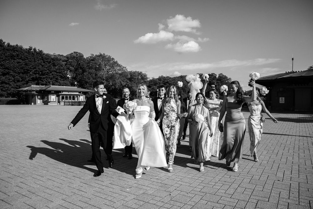 A wedding party walks together on a brick patio on a sunny day with a single puffy cloud in the black and white photo.