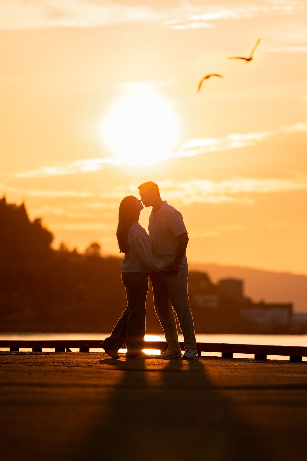 Silhouettes of two figures sitting close together on a railing against a dramatic orange sunset sky.