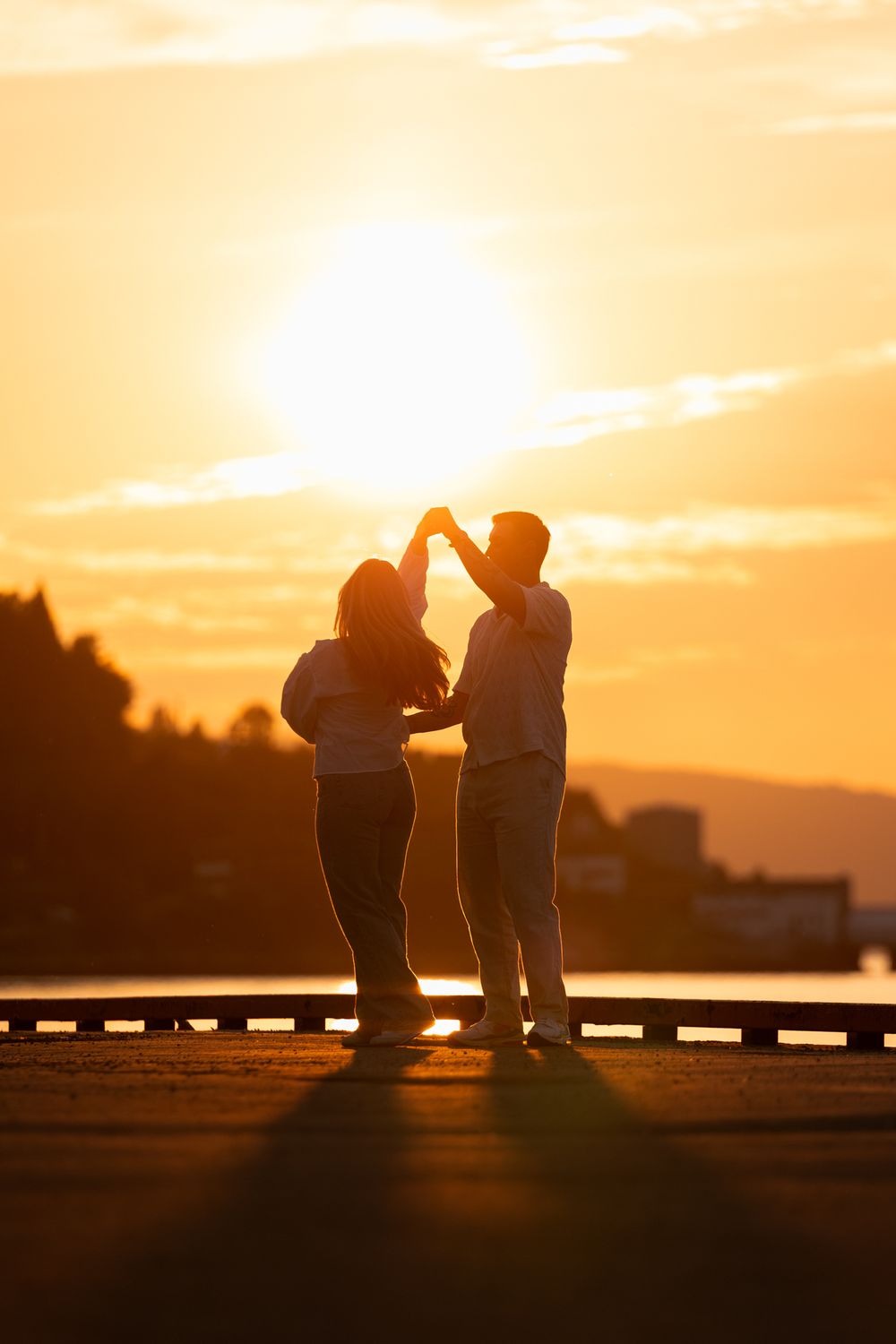 Silhouetted couple dancing together on a pier at sunset with golden sky and mountains in background.