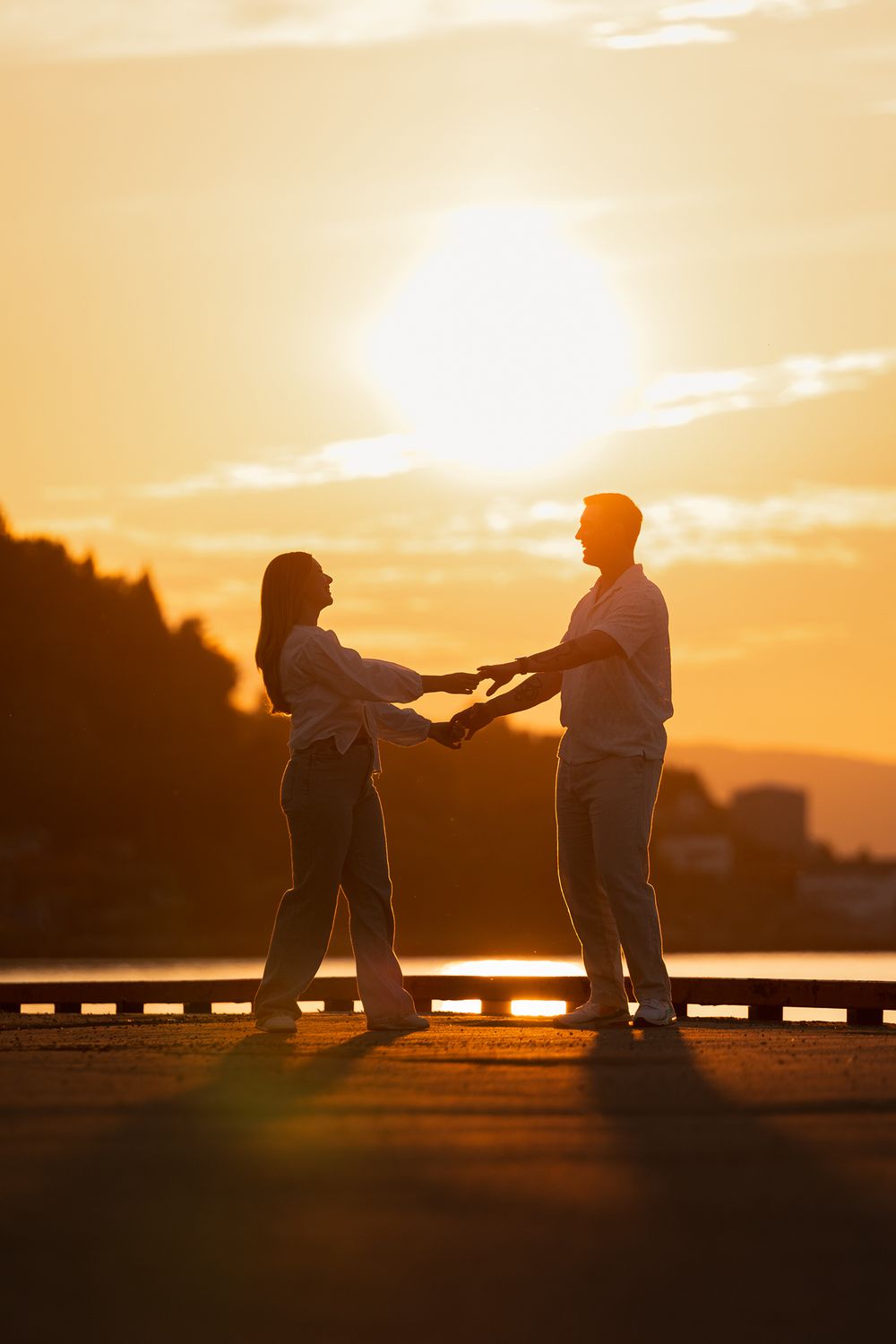 Silhouettes of two people sharing a tender moment at sunset by a waterfront railing.