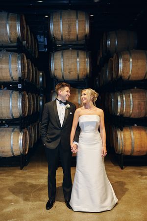 Newlywed couple poses playfully in front of wooden wine barrels in a rustic winery cellar.