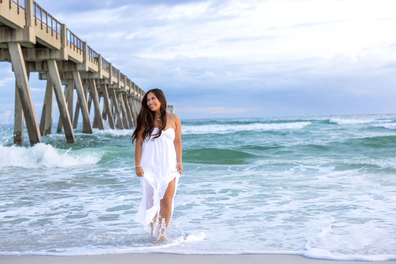 A figure in a flowing white dress stands in the surf beneath a wooden pier as turquoise waves roll onto the beach.