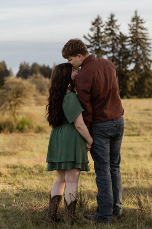 Couple sharing a tender kiss in an open field with evergreen trees, woman in green dress and cowboy boots at scatter creek with photographer