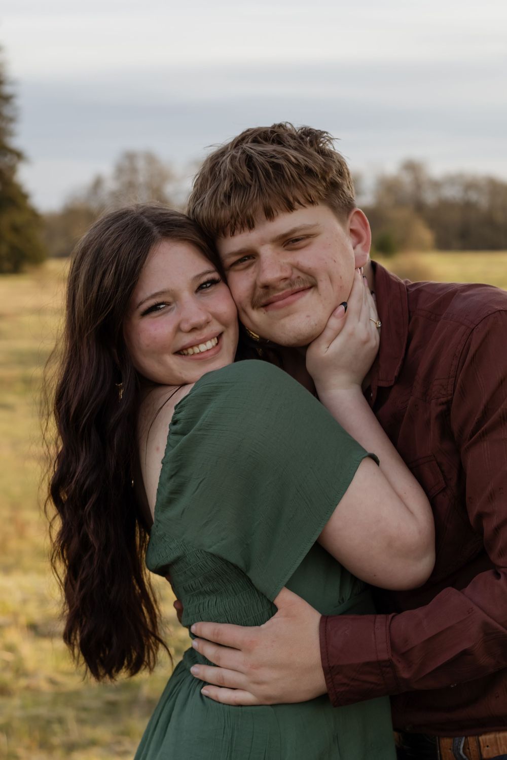 Smiling couple in a warm embrace outdoor, woman in green dress hugging man in burgundy shirt during golden hour photoshoot with photographer