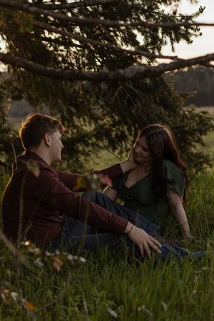 Couple sitting together in lush green grass beneath pine branches during a romantic golden hour photo session in rochester with photographer