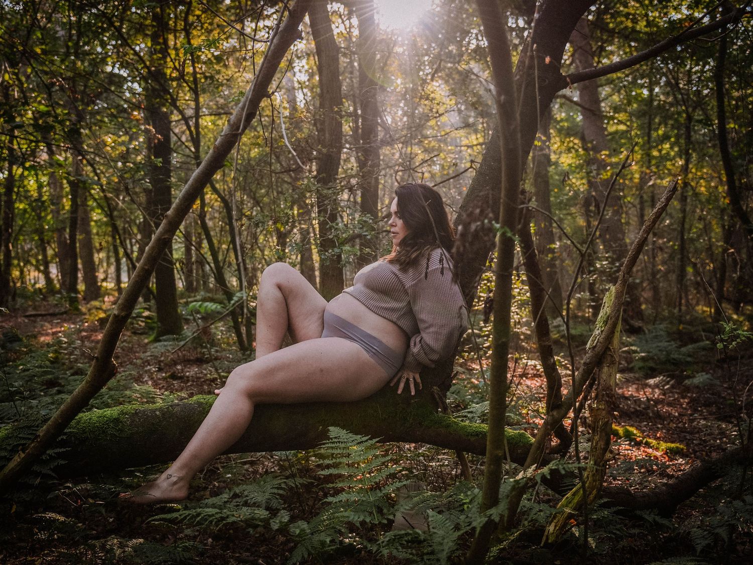 Woman in gray bodysuit relaxing on mossy tree branches in a sunlit forest setting.