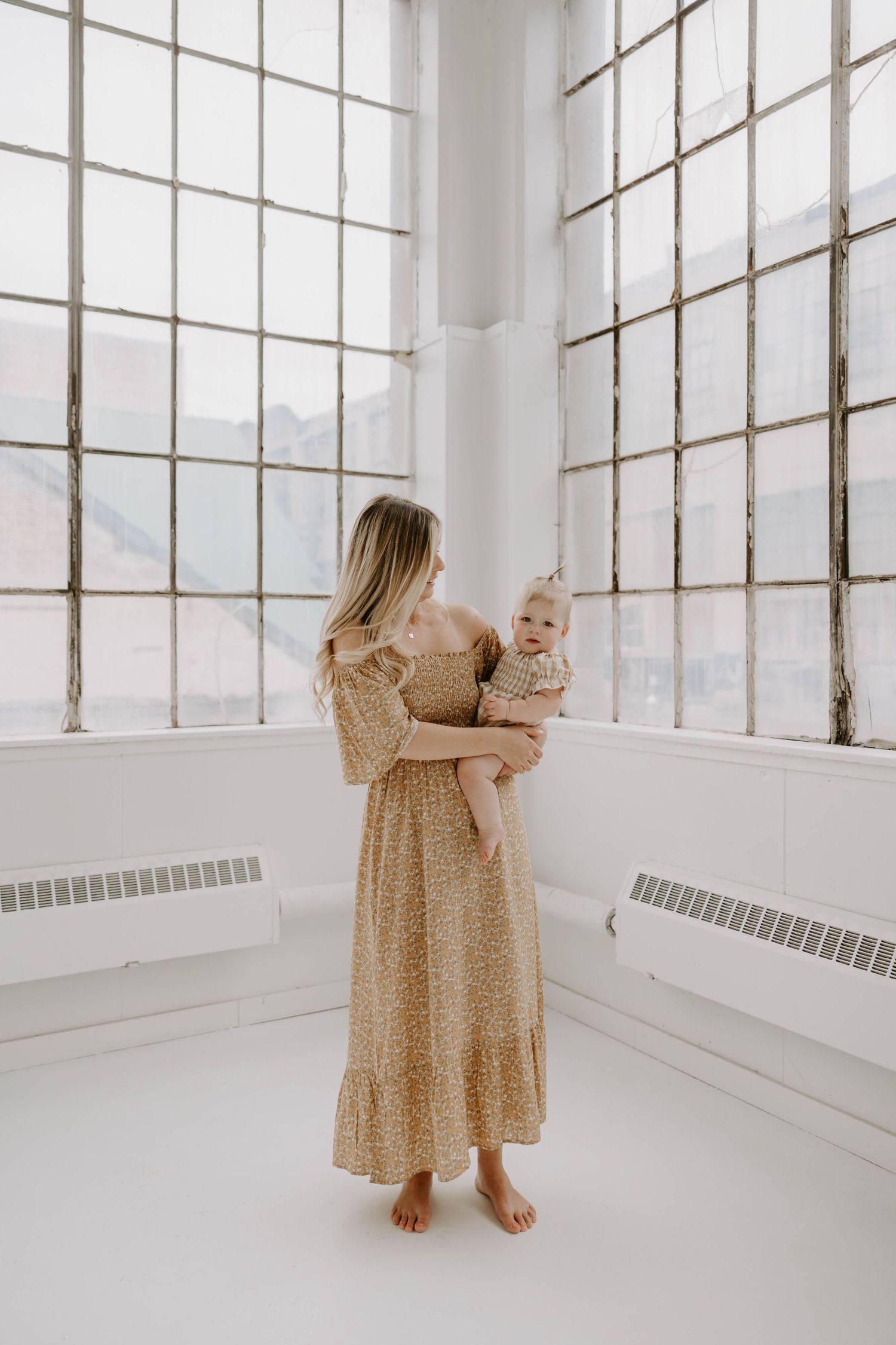 Barefoot mother in floral maxi dress holding baby near large industrial windows.