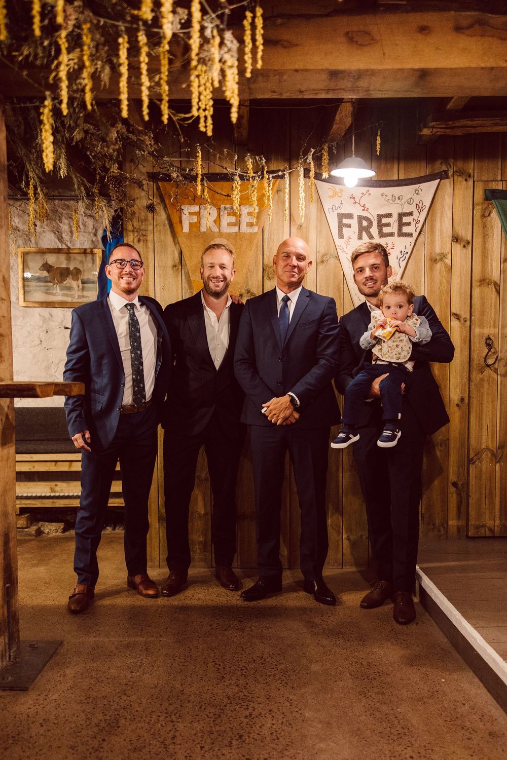 A group in formal attire poses together in a rustic wooden room with string lights overhead.