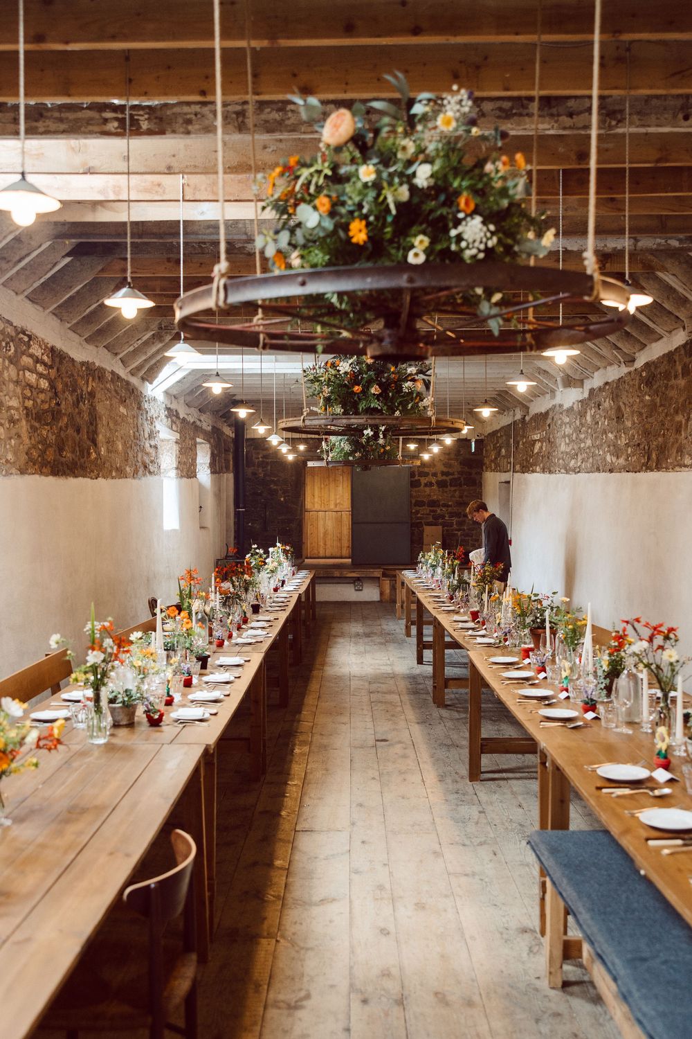 Long rustic dining tables with blue runners and floral arrangements set in a stone-walled venue with overhead lighting.