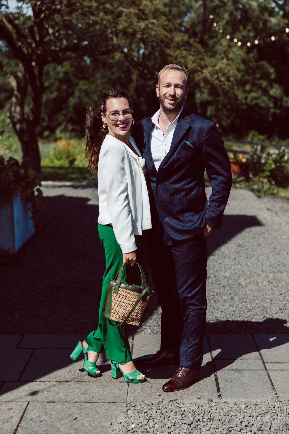 A couple poses together outdoors on stone steps, with one wearing a green skirt and white top and carrying a wicker bag.