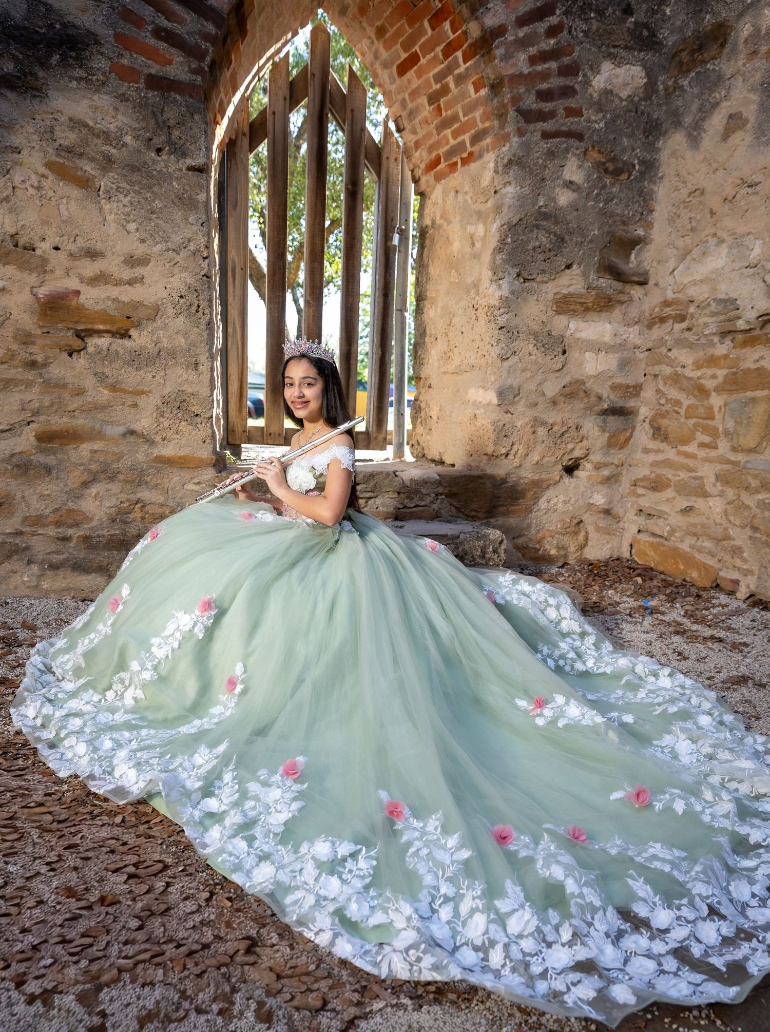 A person in a sparkly purple quinceañera gown with ruffled layers poses against stone pillars outdoors.