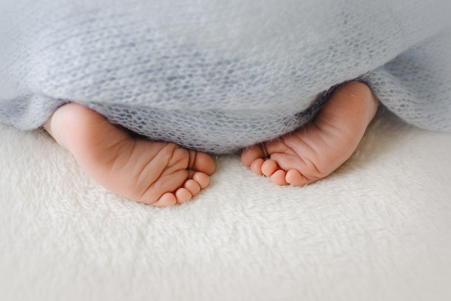 Tiny baby feet peek out from under a soft gray blanket on white carpet.