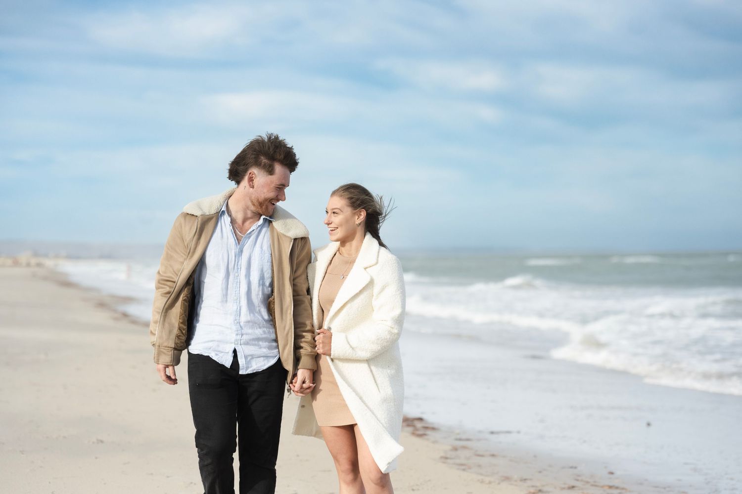 Couple walking hand in hand on a sandy beach, smiling at each other, wearing stylish coats with ocean waves behind.